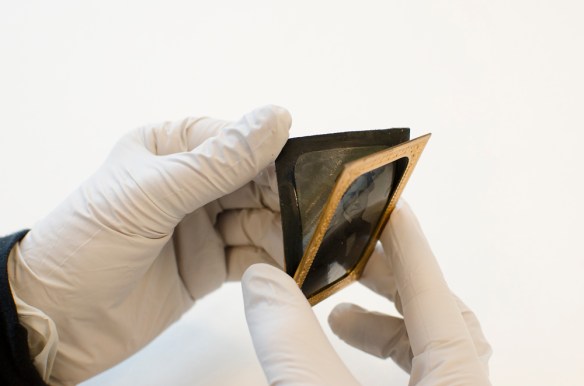 A colour image of gloved hands holding a black-and-white portrait of a man, which is being peeled away from a brass mat with its cover glass.