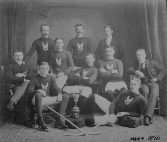 A black-and-white photograph of a group of men standing, sitting on chairs or reclining. Most are wearing the team uniform and hockey skates. 