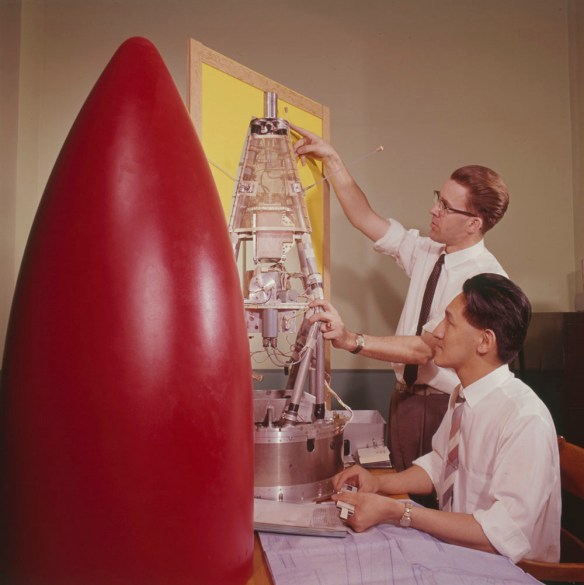 A colour photograph of the red nose cone of a rocket next to two men working on the instruments that will go inside it.