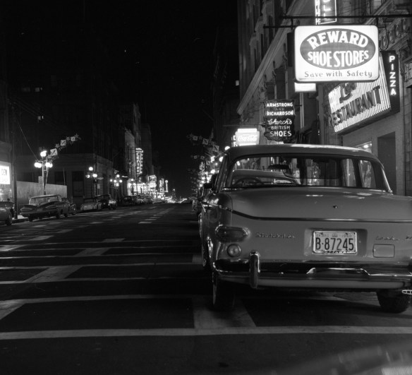 A black-and-white photograph of a street at night with cars parked on both sides and neon store signs adding light.