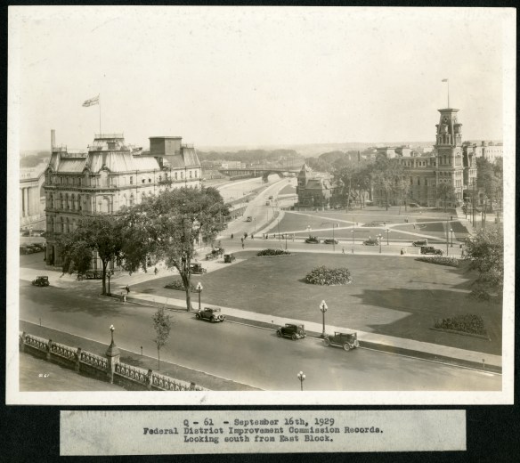 A black-and-white photograph of a quiet park and streets surrounded by two major buildings flying the Union Jack flag from their highest rooftop. Old cars are parked on the main street in the foreground.