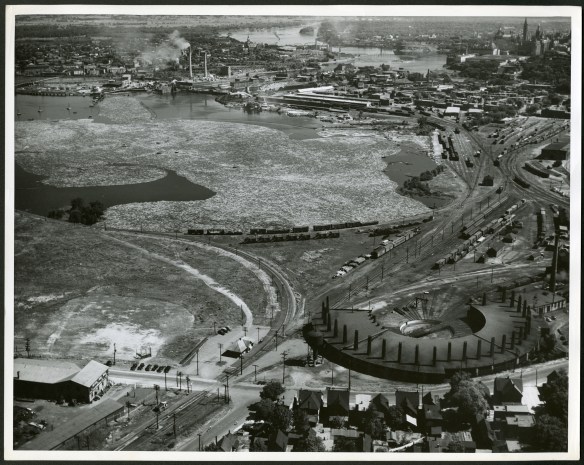 Aerial black-and-white photograph of an industrial landscape with logs floating in the water and a power station and rail lines in the foreground. The Parliamentary Precinct is in the background.