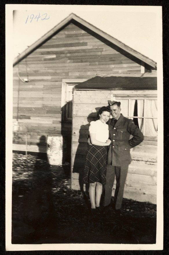 A black-and-white photograph of a man in a military uniform with his arm around a young woman wearing a flowered dress standing in front of a clapboard house.