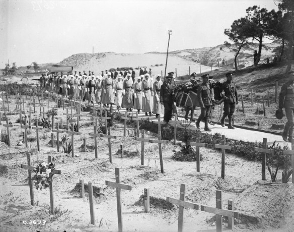 A black-and-white photograph of a funeral procession of soldiers and nursing sisters, accompanying a wheeled stretcher carrier with a flag-draped casket on it, passing through a large cemetery of temporary grave crosses.