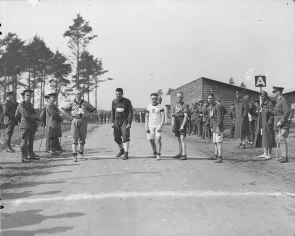 A black-and-white photograph of five men lined up on a road ready to start a race. Soldiers stand and watch from both sides of the road. 
