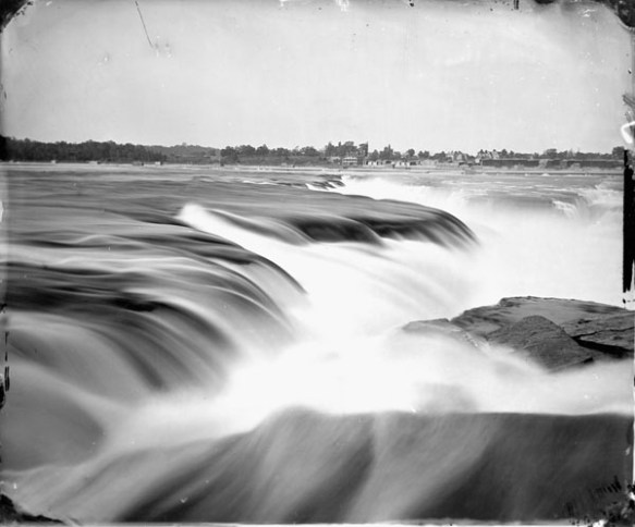 A black-and-white close-up photograph of Chaudière Falls with buildings visible on the distant shore. 