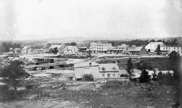 A black-and-white photograph of a sparsely settled town with a few buildings in the background. 