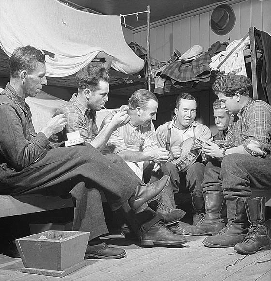 A black-and-white photograph of a group of men sitting around in a bunkroom playing music and smoking.