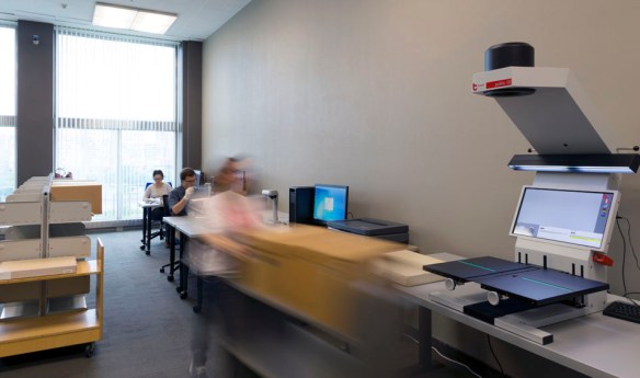 A colour photograph of a room containing a large-format scanner on a table in the foreground, a series of shelving on the left side, and two people sitting at workstations in the background.