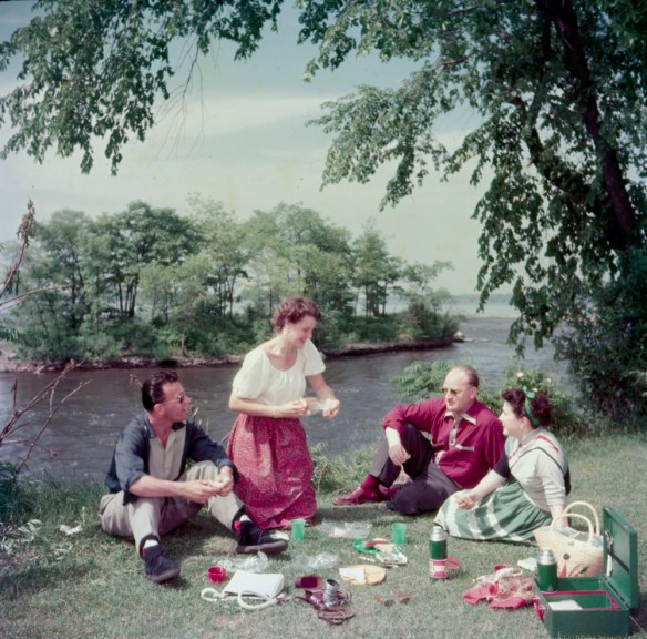 A colour photograph of two couples picnicking next to a river.