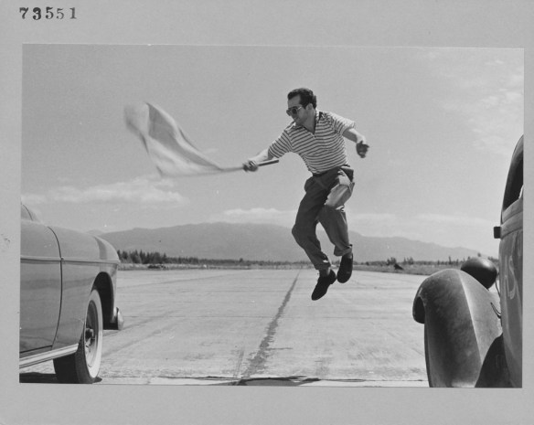 A black-and-white photograph of a man jumping up into the air between two cars, and waving a flag to start a race. 
