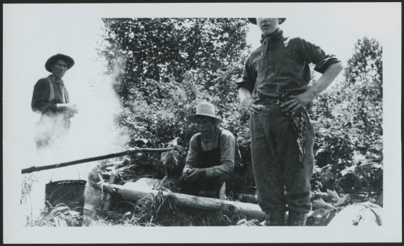 A black-and-white photograph of three men gathered around a fire, presumably having a midday food break. 