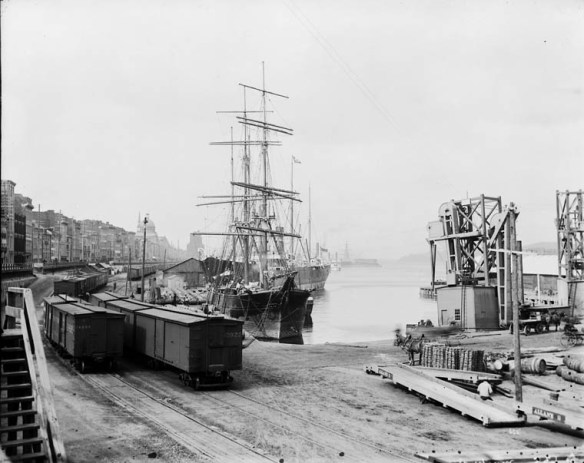 A black-and-white photograph of railway lines running along a ship-lined harbour front.