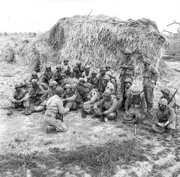 A black-and-white photograph of some two dozen soldiers sitting behind a large hay bale. The soldiers are being briefed before setting out on patrol. 
