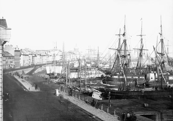 A black-and-white photograph of a busy harbour front, showing a street of buildings and boat-lined piers.