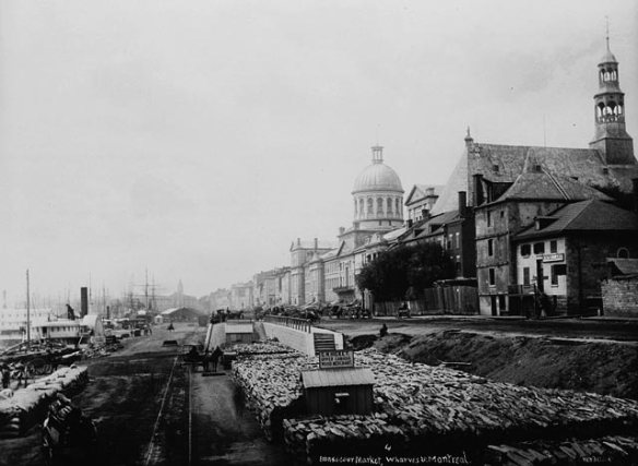 A black-and-white photograph of a wharf lined with various types of cargo with a large neo-classical building and a church along the shoreline.
