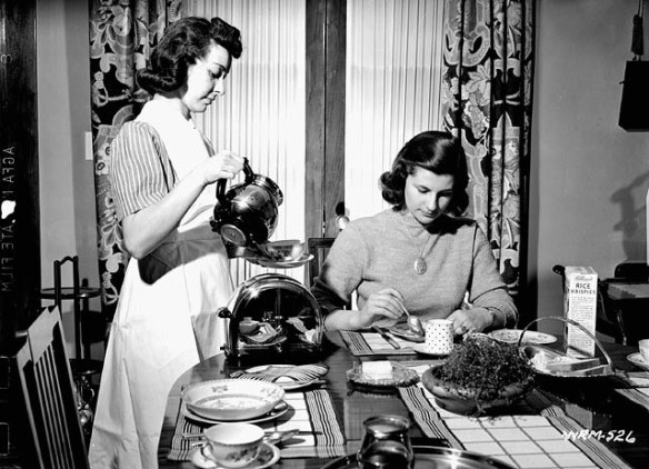 A black-and-white photograph of a standing woman pouring a cup of coffee for another woman sitting at a table eating breakfast.