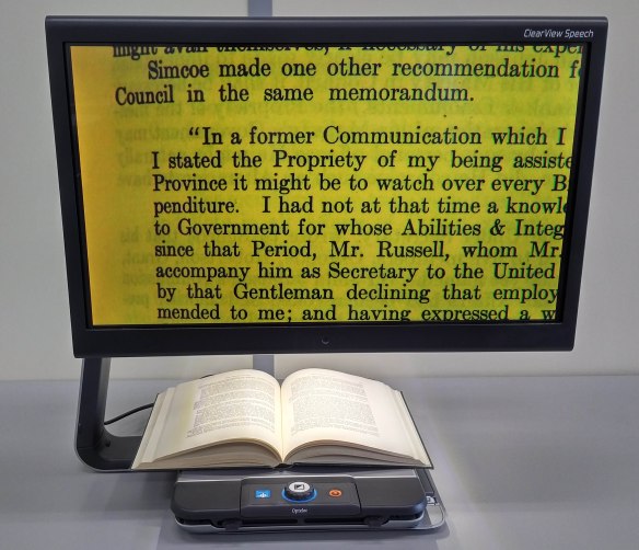 Colour photo of an assistive device having a moving tray with control buttons and an opened book laying on it. The monitor enlarges and displays the book text using a large black font on a yellow background.