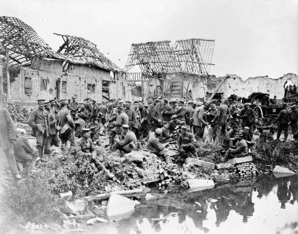 A black-and-white photograph showing a large group of German soldiers milling around between a village and a river or canal. The buildings in the background are mostly destroyed.