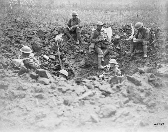 A black-and-white photograph of six soldiers wearing helmets sitting in a large hole in the mud. Some are eating, while others are holding guns and facing away from the camera.