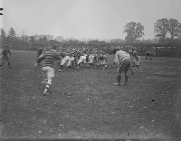 A black-and-white photograph of two rugby teams in a scrum for the ball. Two referees watch the play unfold. 