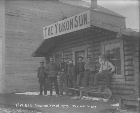 A black-and-white photograph of a group of men standing in front of a log building with a sign that reads The Yukon Sun.