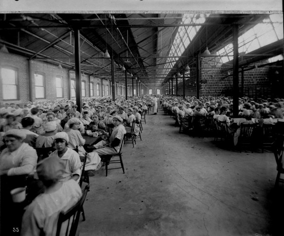 A black-and-white photograph of a large factory dining area seating hundreds of women wearing factory uniforms, seated for their lunchtime meal.