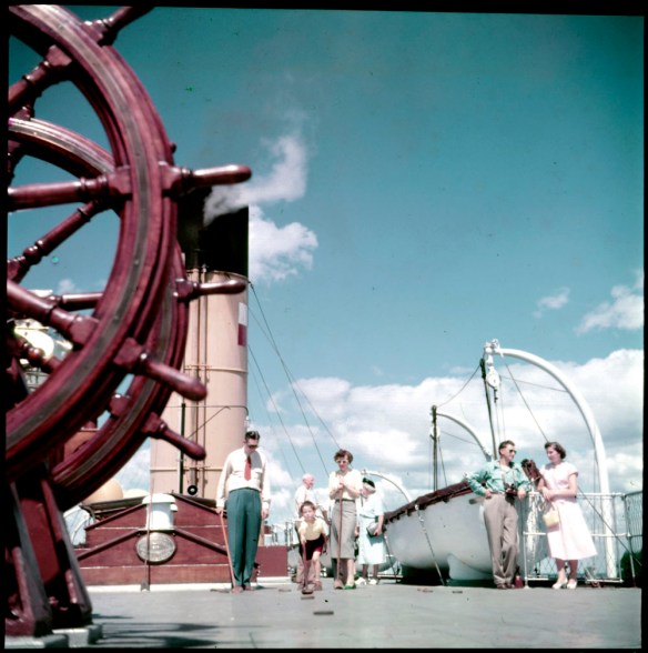 A colour photograph of a boy playing shuffleboard, watched by a man and a woman on the Canadian Pacific Railway cruise ship Assiniboia.