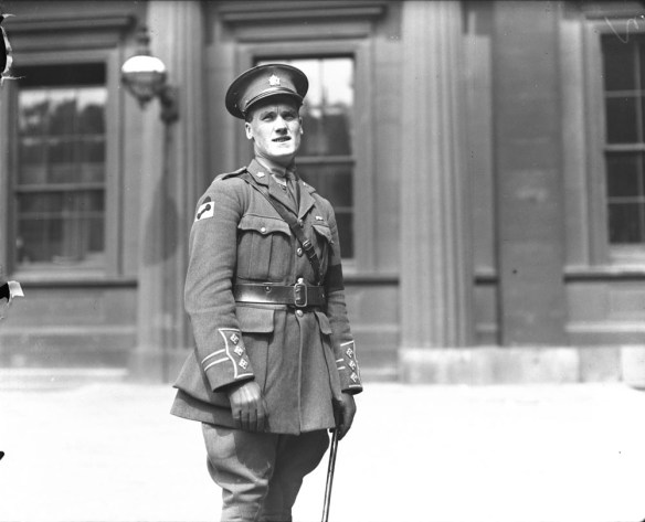 A black-and-white photograph of a soldier in service dress uniform, squinting in the sun and turned slightly away from the camera.