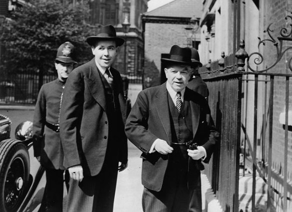 A black-and-white photograph of two men standing in front of an iron gate, with a London police officer behind them to the left.