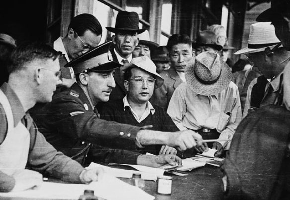 A black-and-white photograph of a Royal Canadian Mounted Police officer seated at a table examining papers with many men around him