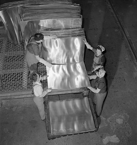 A black-and-white photograph of four women working together to stack square aluminum sheets onto a pallet. 
