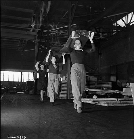 A black-and-white photograph of three women working in unison to carry a long sheet of aluminum over their heads to the inspection table. 