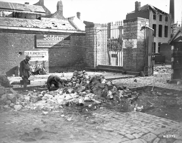 A black-and-white photograph of a town with damaged buildings, with stone and rubble heaped in the middle. One soldier is bent to the ground beside a large pile of debris. A nearby soldier standing to the left is watching him.