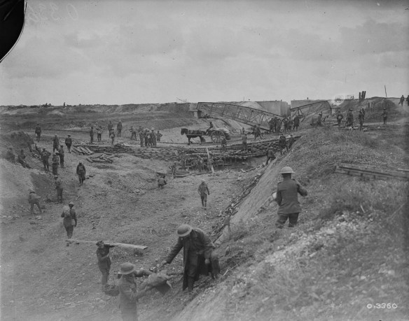 A black-and-white photograph of a dried-out canal with a crooked bridge in the background. A bridge in the middle distance has a horse-drawn carriage crossing it with supplies. Throughout the photo, soldiers are moving about, and some are carrying supplies.