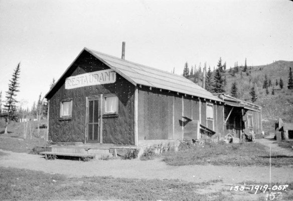 A black-and-white photograph of the exterior of a restaurant located on a dirt road in a remote area.