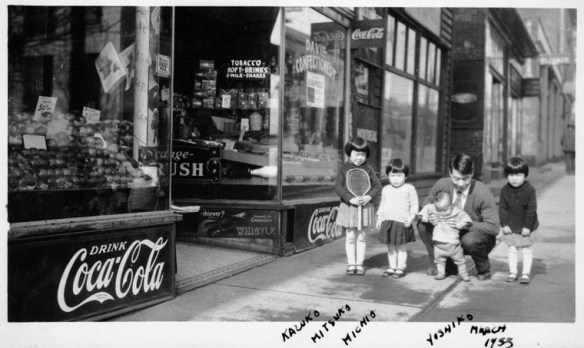 A black-and-white photograph showing a Japanese-Canadian man, who is crouching, and four children in front of a store.