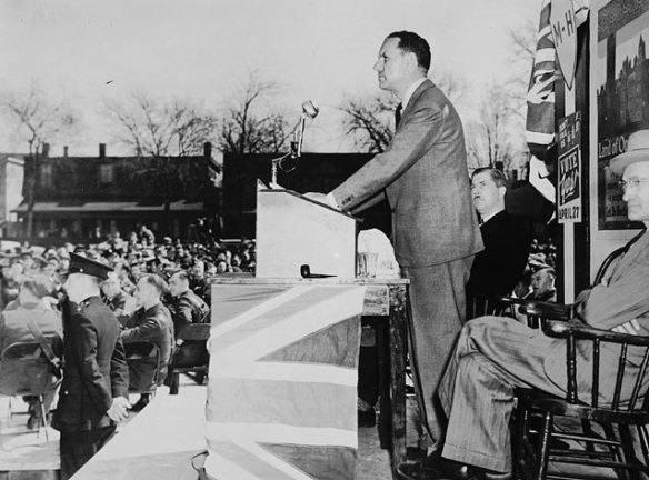 A black-and-white photograph of a man standing at a lectern that is wrapped in the Union Jack, in front of a large audience of workers. Policemen are in the first row.