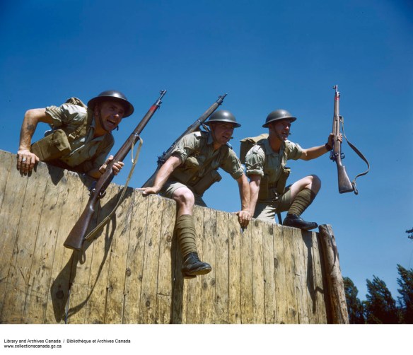 A colour photograph of three men climbing over a wooden fence clad in helmets, short-sleeved shirts, shorts, and socks and boots, and carrying rifles.