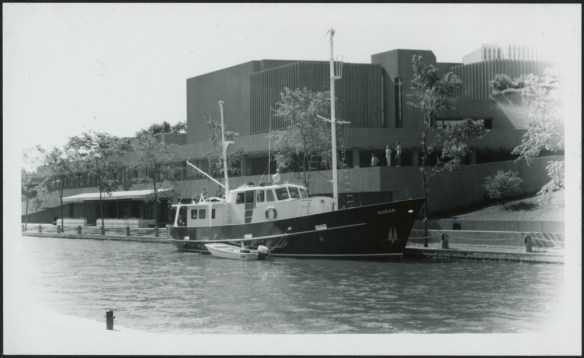 A black-and-white photograph of a moored leisure vessel on a canal beside a large building. 