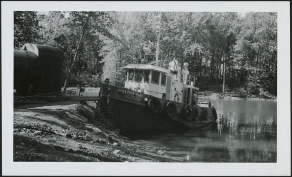 A black-and-white photograph of a small moored fire boat in a wooded stretch of waterway