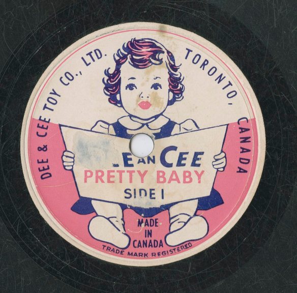 A colour image of a record label for the Dee & Cee Toy Company, Ltd. Side 1 depicts a small girl sitting and holding an open book. The company name and the recording title “Pretty Baby” are on the book cover. 
