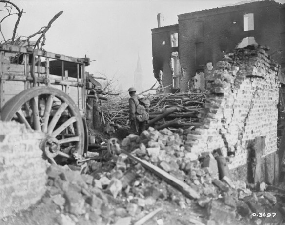 A black-and-white photograph of a uniformed soldier, facing away from the camera and standing in the middle of the bricks and rubble of a destroyed building. A cart with a large wheel is on the left.