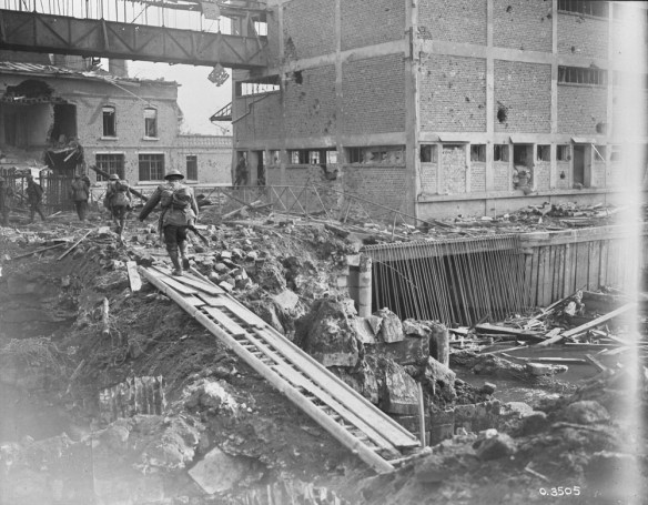 A black-and-white photograph of abandoned buildings, crumbling and filled with holes. The ground is covered in debris, mud, and rocks. Four soldiers are walking away from the camera, having crossed an expanse on a bridge made of debris.
