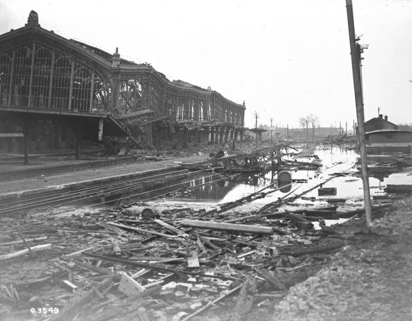 A black-and-white photograph of an ornate railway station in the background, crumpled and heaving, with broken windows and missing walls. The ground is cut diagonally with railway tracks, flooded with water, and littered with debris, planks of wood, and assorted rubble.