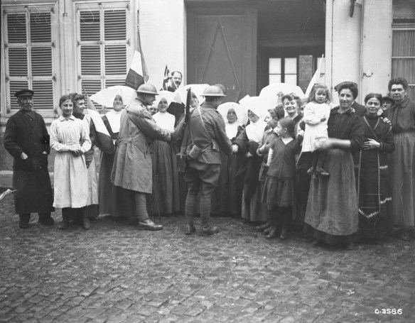 A black-and-white photograph of a group of people standing in front of a building with an open door. Women wearing wide white hats are shaking the hands of soldiers in uniforms. On both ends of the photograph, groups of civilian men and women are smiling into the camera. Someone in the back of the group is holding the French flag.