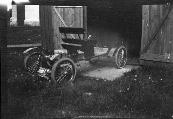 A black-and-white photograph of an early automobile parked outside of a barn. 