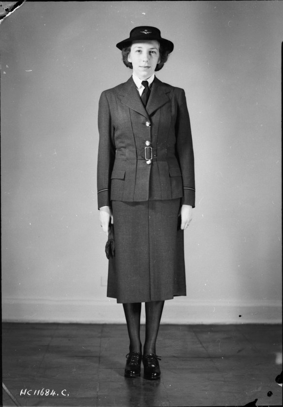 A black-and-white photograph of a woman standing straight to model a military uniform consisting of a Norfolk jacket, skirt, and black shoes. 
