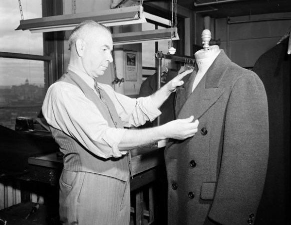 A black-and-white photograph of a tailor adjusting a suit jacket on a tailor’s mannequin. 
