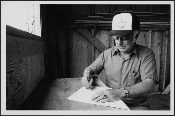 A black-and-white photograph of a man sitting at a rough desk with paper and pen in hand.